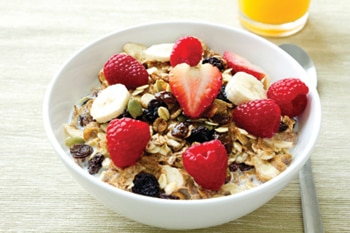 Bowl of whole-grain cereal in milk with fresh strawberries, raspberries, bananas, and raisins, with a glass of orange juice to the side.