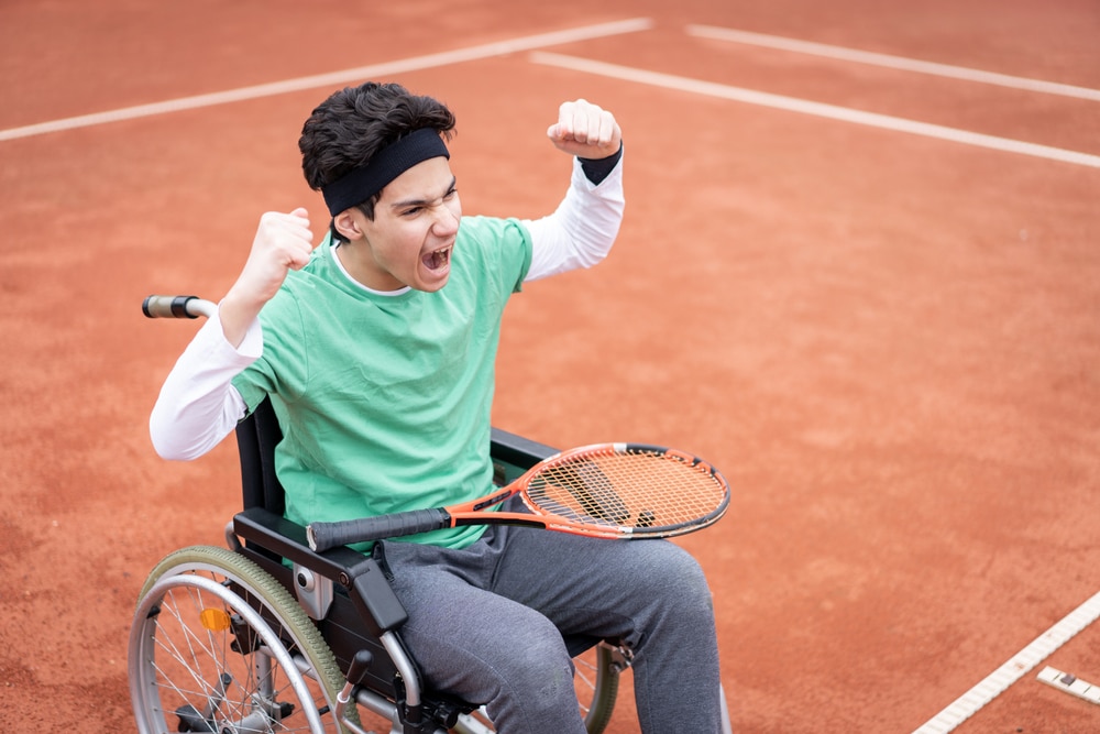 A teen boy in a wheelchair with a tennis racket on his lap and arms raised, celebrating a win on a tennis court.