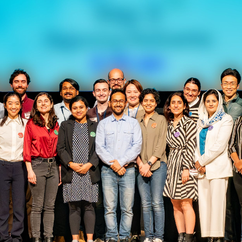 A group of fellows stand on stage in front of a welcome screen, smiling.