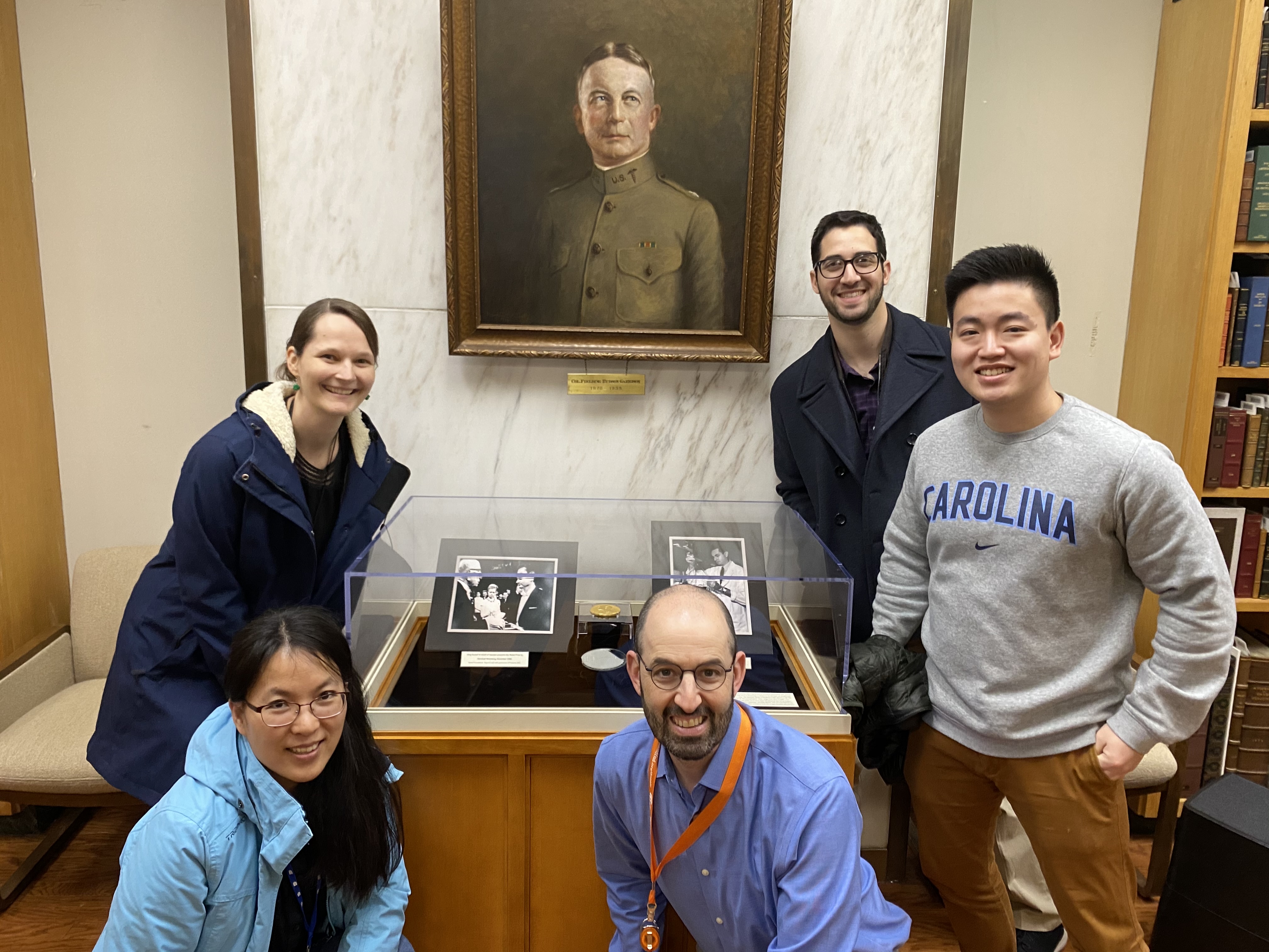 Group picture of 5 people in front of a portrait and a medal in a glass case