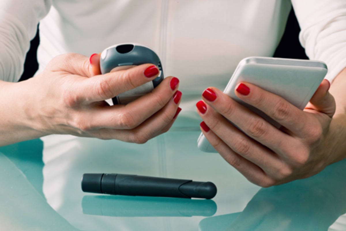 A woman compares her blood glucose reading from a standard glucose monitor with a CGM reading on a smartphone. A lancet used to test blood glucose is on the table in front of her.