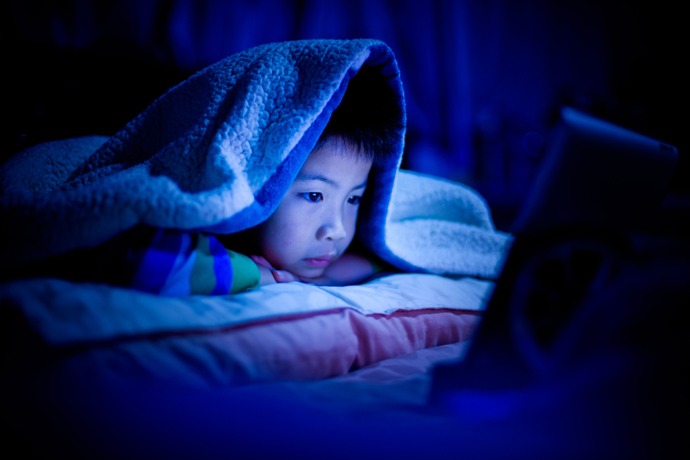 Niño acostado boca abajo en la cama debajo de las cobijas, con las luces apagadas, mira una pantalla que emite luz azul.