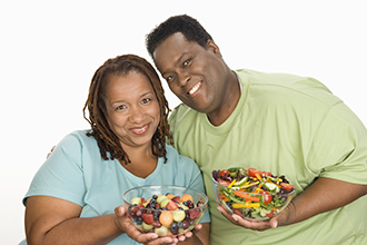 Woman holding a bowl of fruit salad standing next to a man holding a vegetable salad.