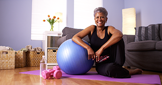 Woman sitting on a yoga mat on her living room floor, leaning on a balance ball, with a pair of weights and water bottle next to her.